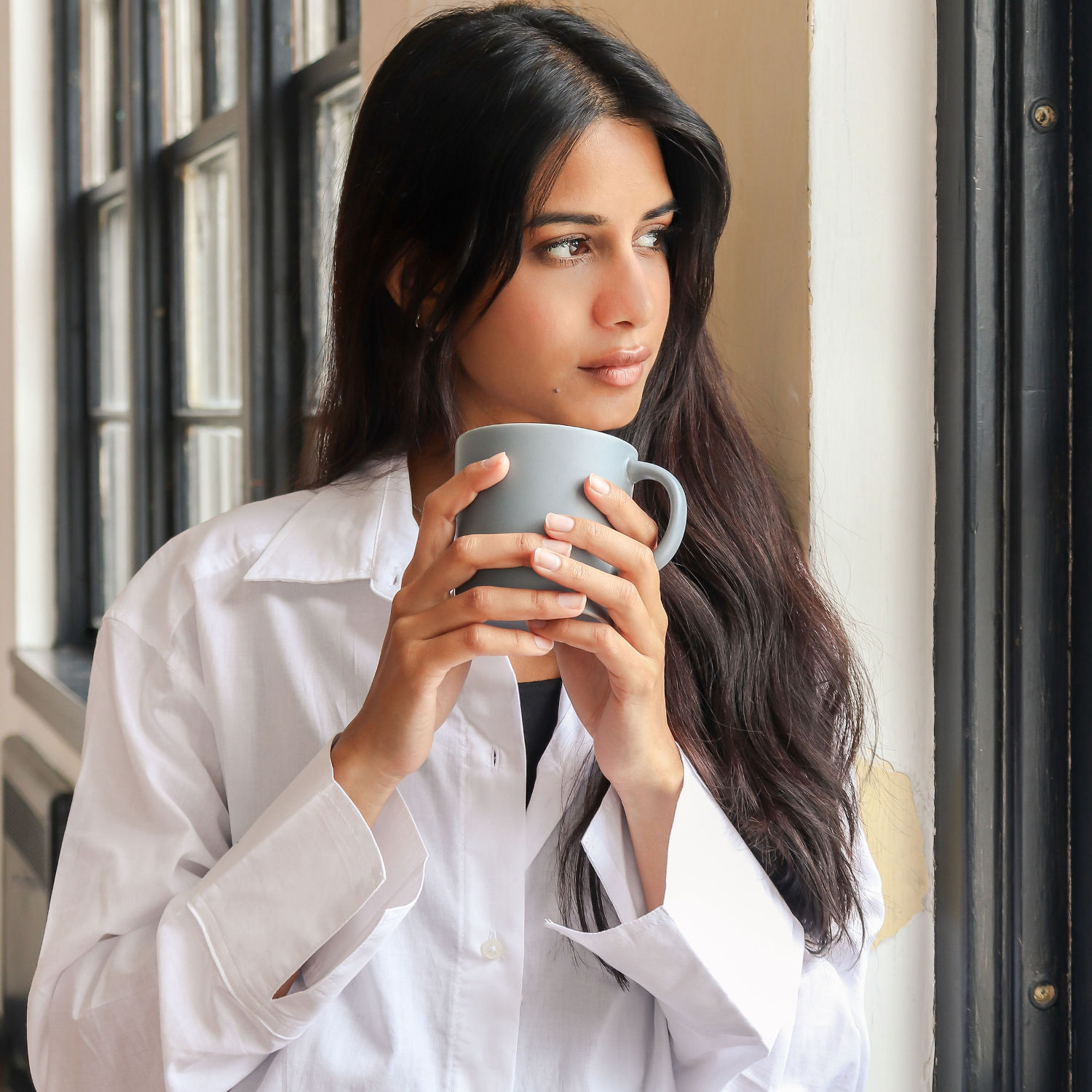 Woman in a white shirt holding a mug by the window, symbolising calm and improved wellbeing through hormone health support